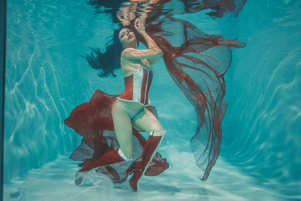 beautiful sexy girl swimming underwater in sporty style red and white fetish corset and over knee thigh high boots with heels - Photo, Image