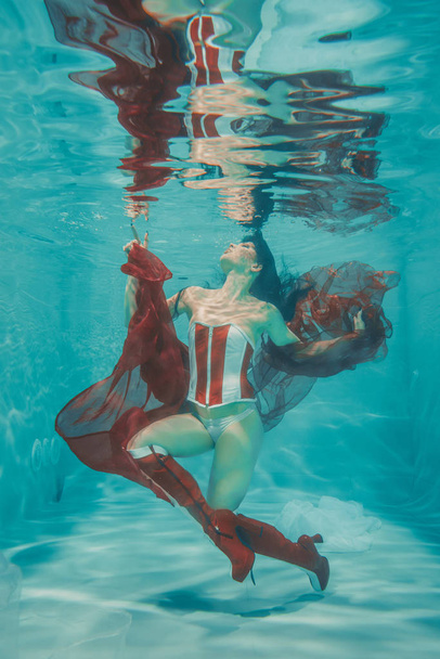 όμορφη σέξι κορίτσι υποβρύχια κολύμβηση σε σπορ στυλ κόκκινο και άσπρο φετίχ κορσέ και πάνω από το γόνατο μπότες μέχρι το μηρό ψηλά με τακούνια - Φωτογραφία, εικόνα