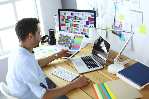 Portret młodego projektanta siedzącego w pracowni graficznej przed laptopem i komputerem podczas pracy online. - Zdjęcie, obraz