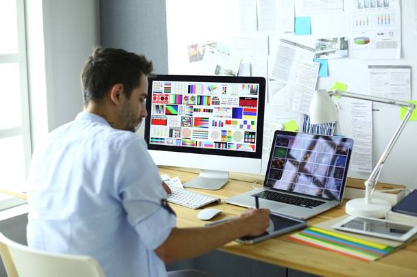 Porträt eines jungen Designers, der im Grafikstudio vor Laptop und Computer sitzt, während er online arbeitet. - Foto, Bild
