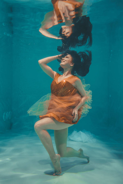 mooi model meisje onder water zwemt in oranje jurk en geniet van de ontspanning en het ontbreken van stress - Foto, afbeelding