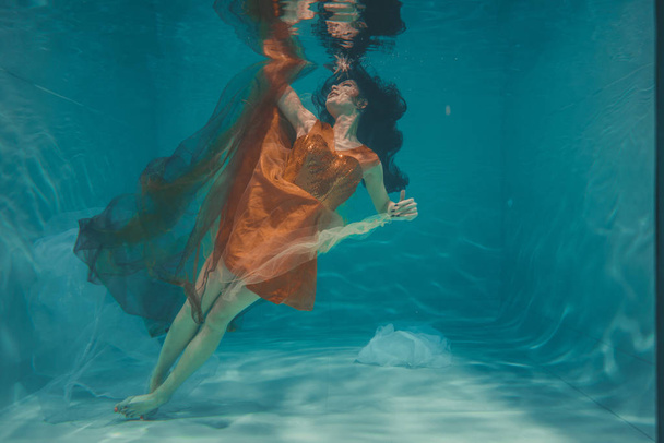 gyönyörű modell lány úszik a víz alatt a narancssárga ruha, és élvezi a relaxációs és stressz hiánya - Fotó, kép