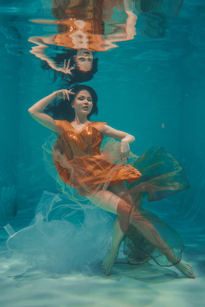 το όμορφο μοντέλο κορίτσι υποβρύχια κολυμπά σε πορτοκαλί φόρεμα και απολαμβάνει την χαλάρωση και την έλλειψη στρες - Φωτογραφία, εικόνα