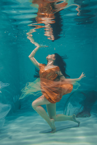 το όμορφο μοντέλο κορίτσι υποβρύχια κολυμπά σε πορτοκαλί φόρεμα και απολαμβάνει την χαλάρωση και την έλλειψη στρες - Φωτογραφία, εικόνα