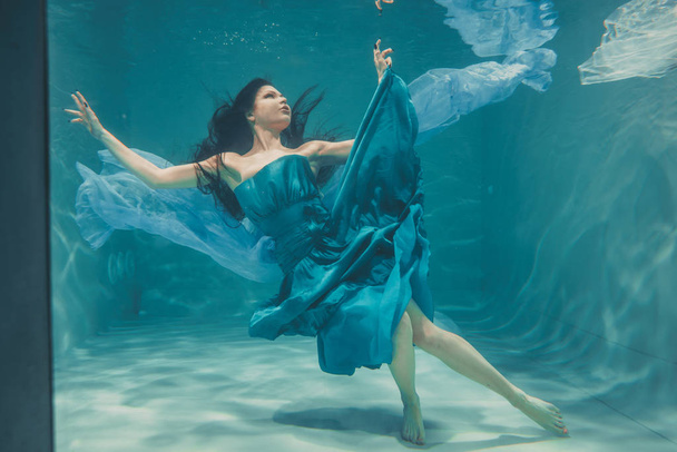 mooi model meisje met lange zwarte haren zwemt onder water in avond blauwe jurk en geniet van de ontspanning en het ontbreken van stress - Foto, afbeelding