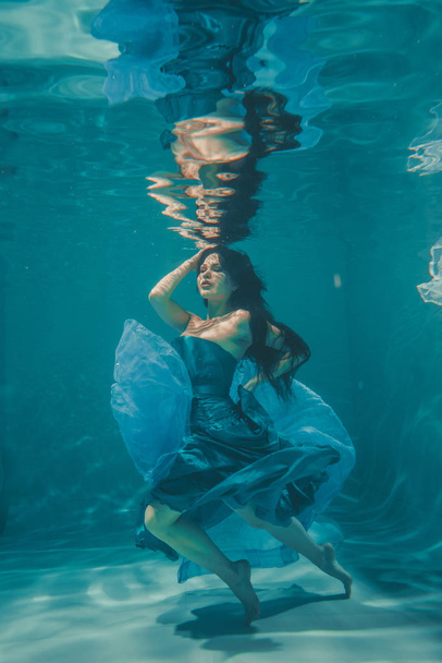 mooi model meisje met lange zwarte haren zwemt onder water in avond blauwe jurk en geniet van de ontspanning en het ontbreken van stress - Foto, afbeelding