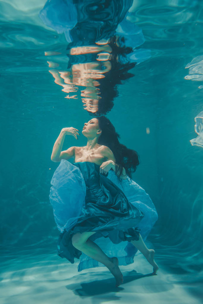το όμορφο μοντέλο κορίτσι με μακριά μαύρα μαλλιά υποβρύχια κολυμπά βράδυ μπλε φόρεμα και απολαμβάνει την χαλάρωση και την έλλειψη στρες - Φωτογραφία, εικόνα