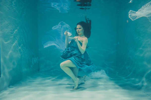 funny girl having fun and drinking soothing tea for Breakfast while meditatively swimming underwater in a dress - Photo, Image