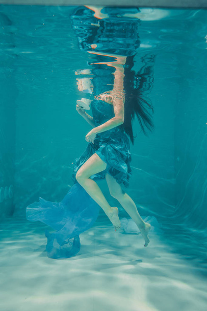 grappig meisje met plezier en rustgevende thee drinken voor het ontbijt terwijl meditatively onderwater zwemmen in een jurk - Foto, afbeelding