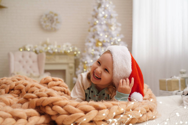 designer blankets from Merino beige! laughing child in Santa hat. behind the girl in the background is a Christmas tree by the window. happy child lying on the bed rejoicing at the onset of Christmas - 写真・画像