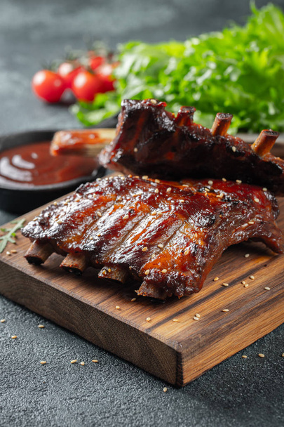 Closeup of pork ribs grilled with BBQ sauce and caramelized in honey. Tasty snack to beer on a wooden Board for filing on dark concrete background - Foto, Imagen