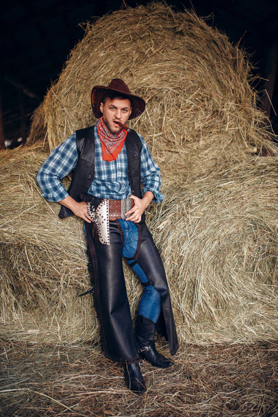 Brutale cowboy vormt met sigaar en revolver, hooiberg op westerse achtergrond. Vintage mannelijke persoon met pistool op boerderij, wild west cultuur - Foto, afbeelding