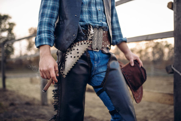 Ковбой в кожаной одежде позирует с сигарой в лошадином загоне, ранчо Техаса, вестерн. Винтажный мужчина на ферме, образ жизни дикого запада
 - Фото, изображение