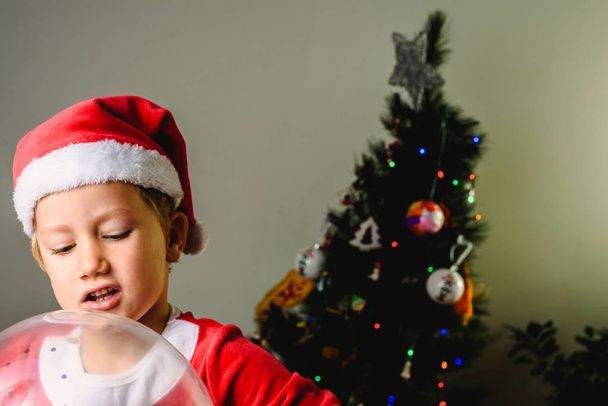 Portrait of blond and handsome 5 year old boy as Santa Claus in the foreground playing with a balloon and christmas tree on white background. - Photo, Image