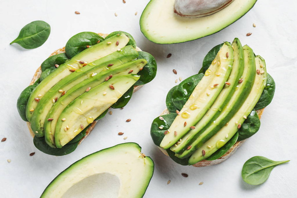 Twee ciabatta toast met gesneden avocado olijf olie, spinazie en vlas en sesam zaadjes. Gezond vegetarisch ontbijt op witte achtergrond. Bovenaanzicht. Plat leggen - Foto, afbeelding