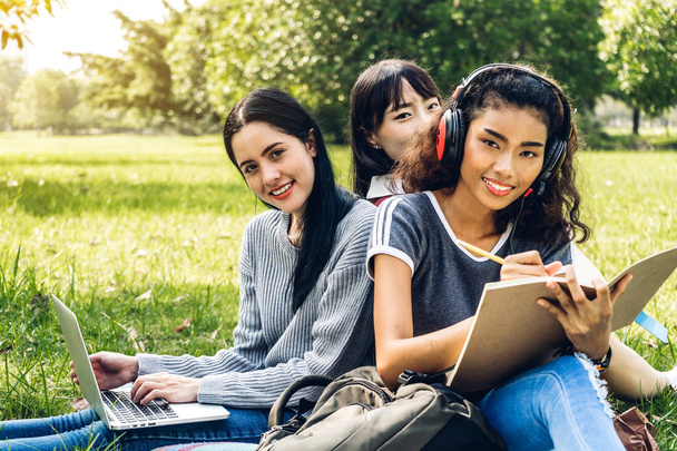 Grupo de estudiantes internacionales sonrientes o adolescentes sentados y usando computadora portátil haciendo sus deberes con libro juntos en el parque en la universidad. - Foto, imagen