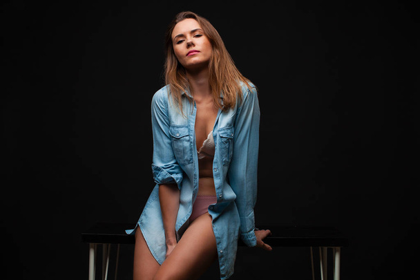 Portrait of a young sexy girl in a denim shirt and nude beautiful legs sitting on a table in a dark studio - Foto, Imagem