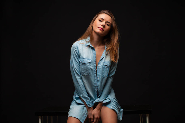 Portrait of a young sexy girl in a denim shirt and nude beautiful legs sitting on a table in a dark studio - 写真・画像