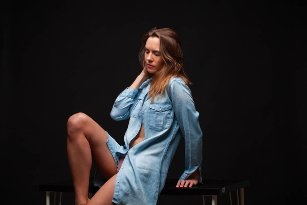 Portrait of a young sexy girl in a denim shirt and nude beautiful legs sitting on a table in a dark studio - Fotografie, Obrázek