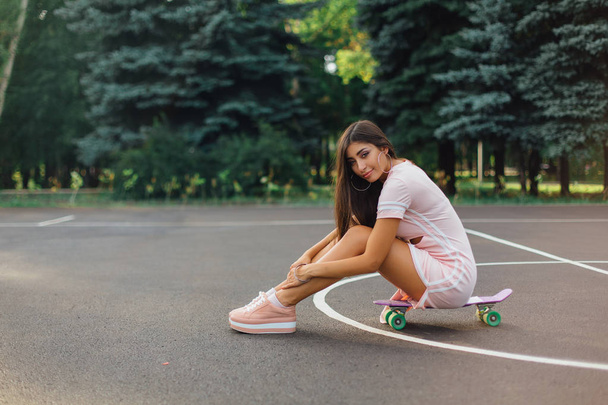 Portrait of a smiling charming brunette female sitting on her skateboard on a basketball court. - Zdjęcie, obraz