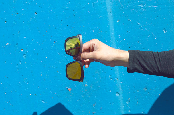 Ο άνθρωπος κρατώντας στα χέρια ένα στιλάτα γυαλιά ηλίου πάνω σε μπλε φόντο. - Φωτογραφία, εικόνα