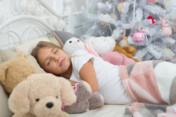 Enjoying xmas at home. Little child sleep at Christmas tree. Little girl lying in bed with toys. Cute girl fell asleep on Christmas night. Sleeping child. Peaceful sleep and rest on winter holidays - Photo, Image