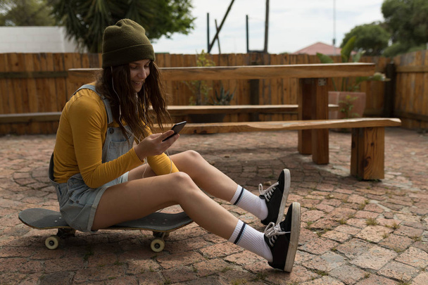 Стильная женщина-скейтбордистка с помощью мобильного телефона, сидя на скейтборде
 - Фото, изображение
