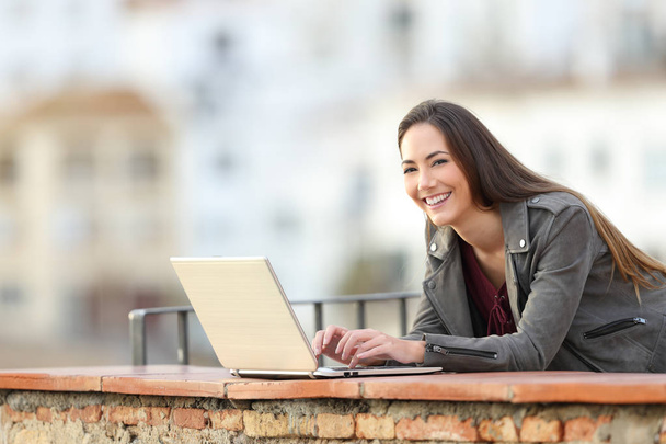 Happy woman using a laptop looking at camera on vacation in a rural apartment terrace - Foto, Imagem