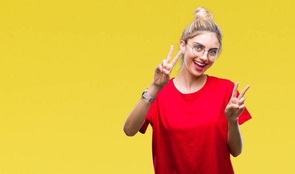 junge schöne blonde Frau mit rotem T-Shirt und Brille vor isoliertem Hintergrund lächelt in die Kamera und zeigt Finger, die Siegeszeichen machen. Nummer zwei. - Foto, Bild