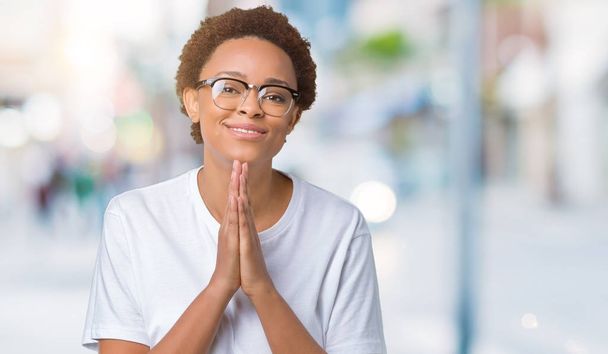 Hermosa mujer afroamericana joven con gafas sobre fondo aislado rezando con las manos juntas pidiendo perdón sonriendo confiado
. - Foto, Imagen