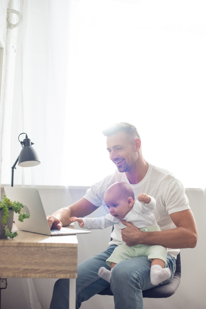 happy father sitting on chair, holding baby daughter and using laptop at home - Foto, Imagem