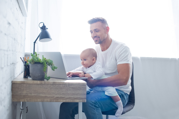smiling father sitting on chair, holding baby and using laptop at home - Photo, Image