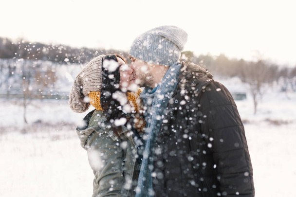 Happy woman and man, kissing, and huging in snowfall, spending time together enjowing eachother, snow, life, winter. Portrait wonderful young couple wearing stylish wool hats and scarfs. - Photo, Image
