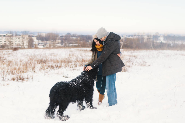 Красивий чоловік і жінка насолоджуються снігом під час прогулянки зі своїм великим чорним кучерявим собакою, обіймаючи один одного і прив'язуючи свого пухнастого друга взимку, з містом і небом на задньому плані
 - Фото, зображення