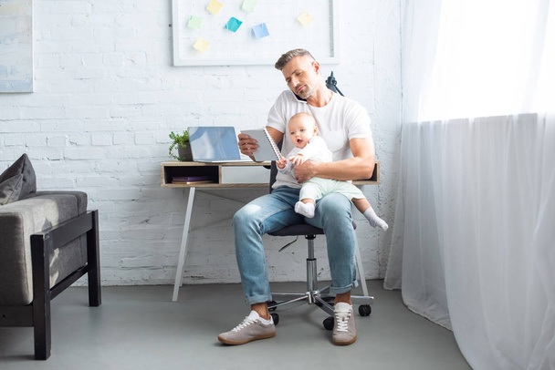 father sitting on chair, looking at notebook, holding adorable baby daughter and talking on smartphone at home - Foto, Bild