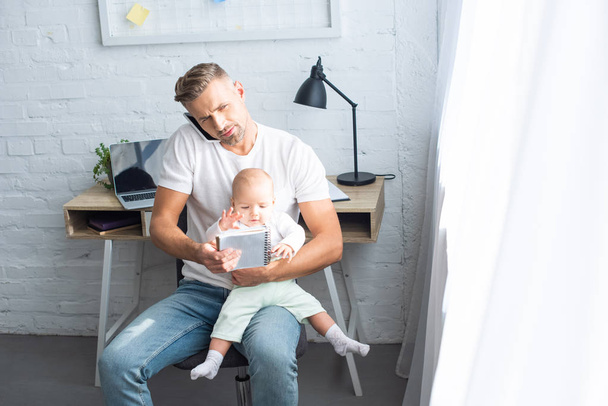 busy father sitting on chair with notebook, talking on smartphone and holding baby daughter at home - Photo, Image