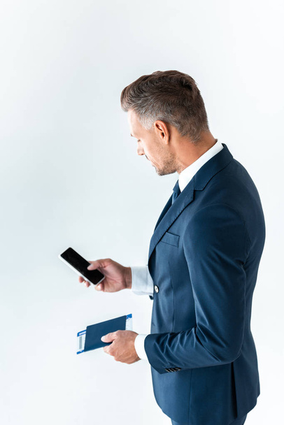 високий кут зору красивого бізнесмена, використовуючи смартфон з порожнім екраном і тримаючи паспорт з квитком ізольовано на білому
 - Фото, зображення