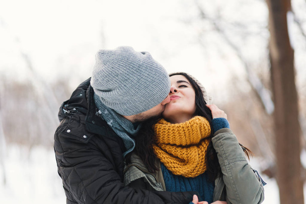 Happy woman and man, kissing, and huging, spending time together enjowing eachother, snow, life, winter. Portrait wonderful young couple wearing stylish wool hats and scarfs. - Фото, зображення