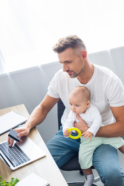 handsome father sitting at computer desk, doing online shopping with credit card and holding baby daughter at home  - Photo, Image