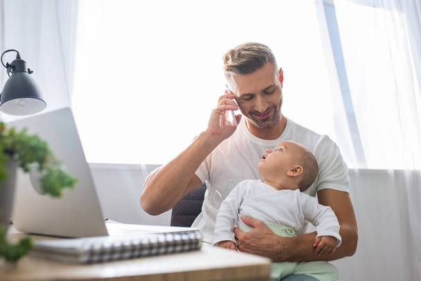 happy father sitting at computer desk, talking on smartphone and holding baby daughter at home - Photo, Image