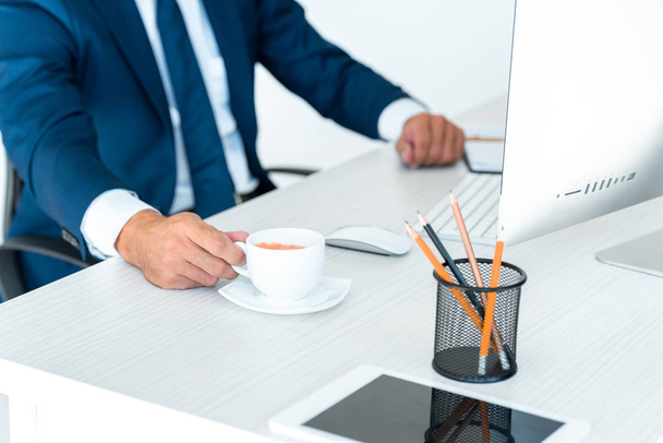 abgeschnittenes Bild eines Geschäftsmannes, der eine Tasse Kaffee am Tisch einnimmt, isoliert auf Weiß - Foto, Bild