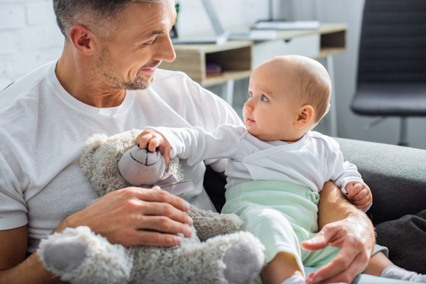 adorable baby daughter playing with teddy bear while sitting with smiling father on couch at home - Foto, Bild