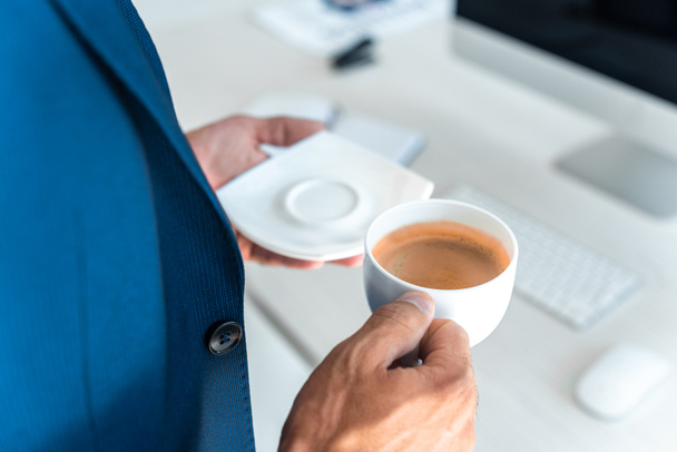 περικομμένη εικόνα του επιχειρηματία κρατώντας φλιτζάνι καφέ και πλάκα στο γραφείο - Φωτογραφία, εικόνα