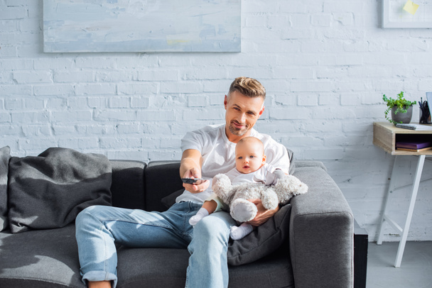 father sitting on couch with adorable baby daughter, holding remote controller and watching television at home - Foto, Bild