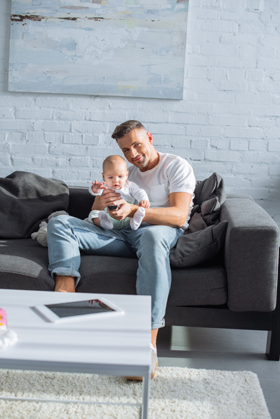 glücklicher Vater, der mit seiner entzückenden kleinen Tochter auf der Couch sitzt und im Wohnzimmer in die Kamera schaut - Foto, Bild