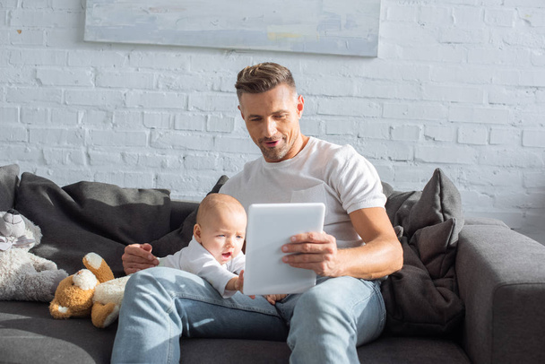 father sitting on couch with baby daughter and using digital tablet in living room - Photo, Image
