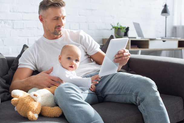 father sitting on couch with adorable baby daughter and using digital tablet at home - Foto, immagini
