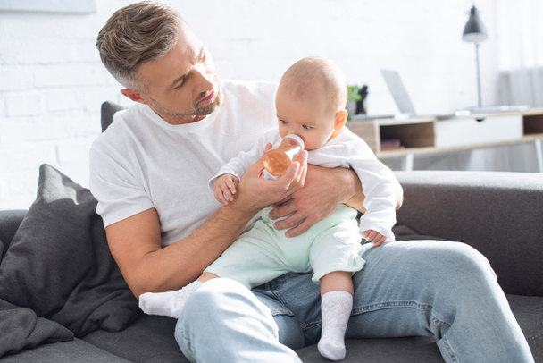 handsome father sitting on couch and feeding baby daughter from bottle at home - Photo, Image