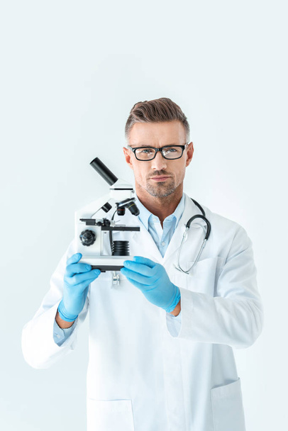 handsome serious scientist in glasses holding microscope and looking at camera isolated on white - Fotografie, Obrázek