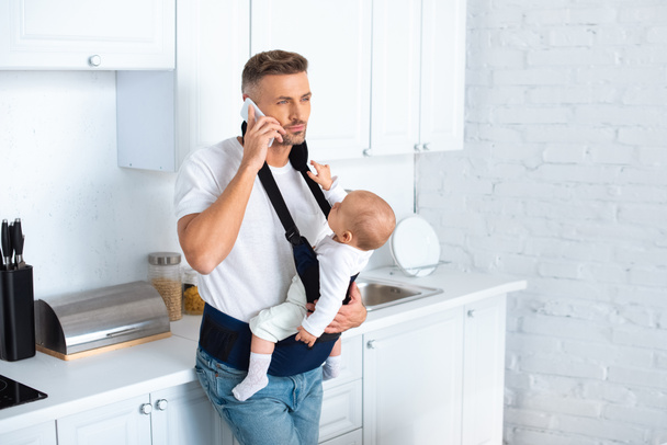 serious father holding infant daughter in baby carrier and talking on smartphone in kitchen - Fotó, kép